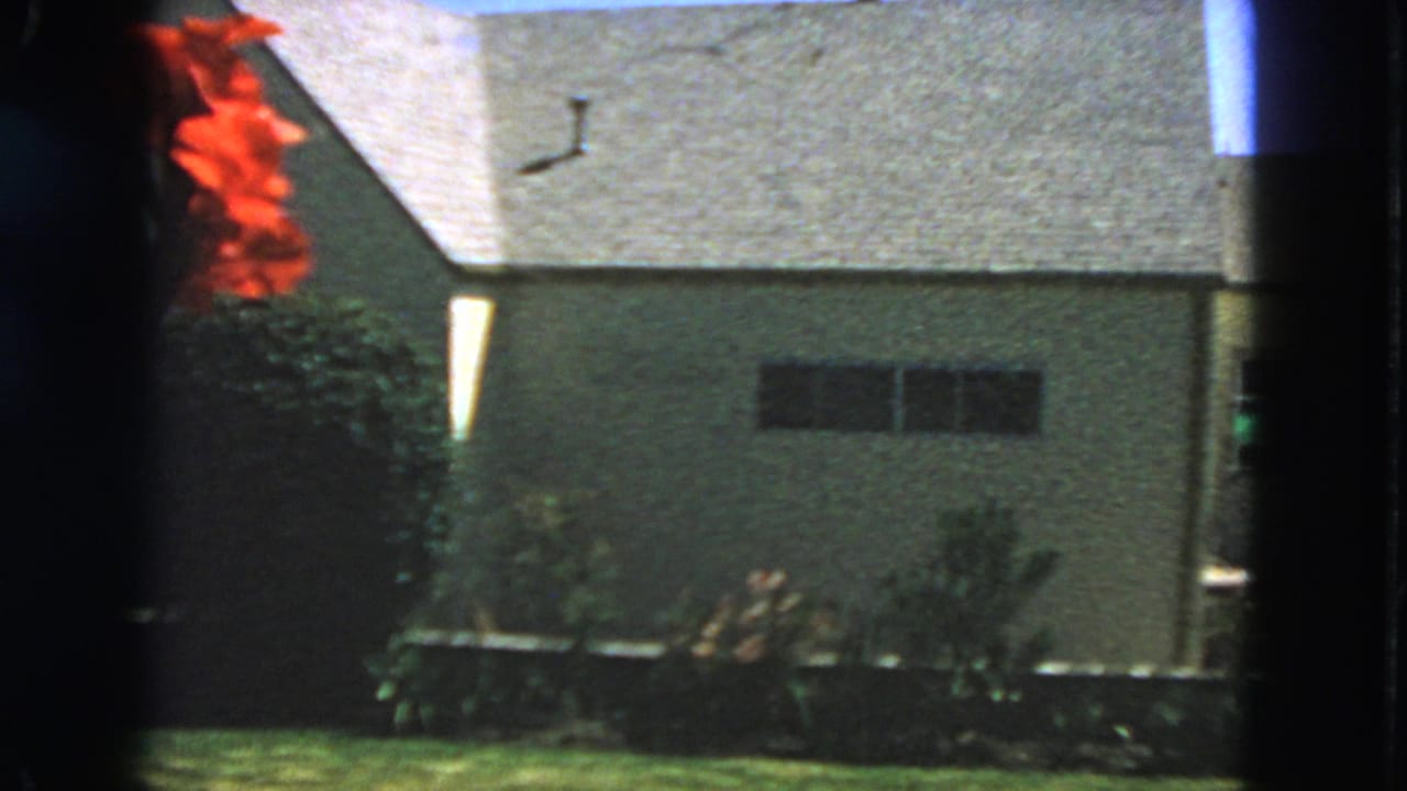 1963 San Francisco home-movie frame showing a light stucco house exterior with roof shingles, a dark window, lawn, shrubs, and red flowers.