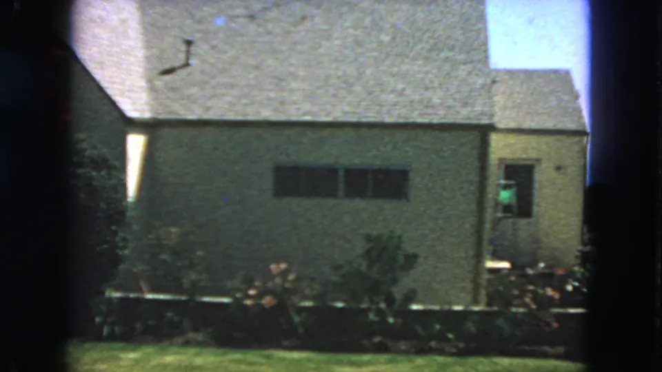 1963 San Francisco frame of a stucco house exterior with a side door, dark window, planted yard, lawn, and blue sky.