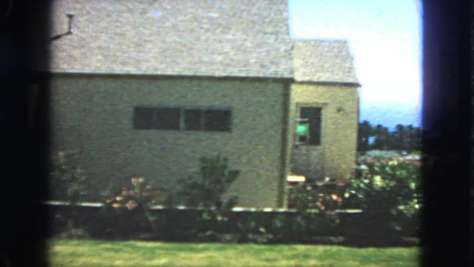 1963 San Francisco home-movie view of a stucco house facade with a dark window, side door, roofline, shrubs, and lawn.