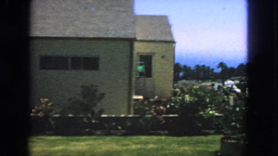 1963 San Francisco frame showing the house side, lawn, shrubs, distant palm trees, blue sky, and dark 8mm frame edges.