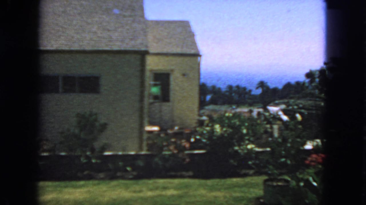 1963 San Francisco home-movie frame showing a light-colored house, lawn, shrubs, and trees through dark window edges.