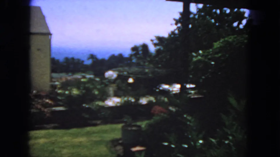 1963 San Francisco 8mm frame with a house wall, planted yard, trees, and bright sky seen from inside or behind a window.