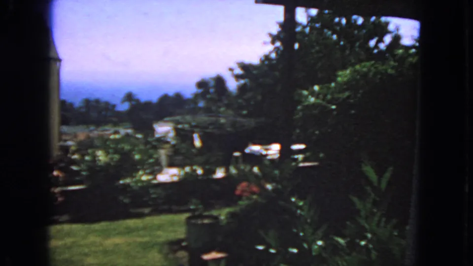 1963 San Francisco home-movie still showing a breezy garden, lawn, dense plants, and a partial residential exterior.