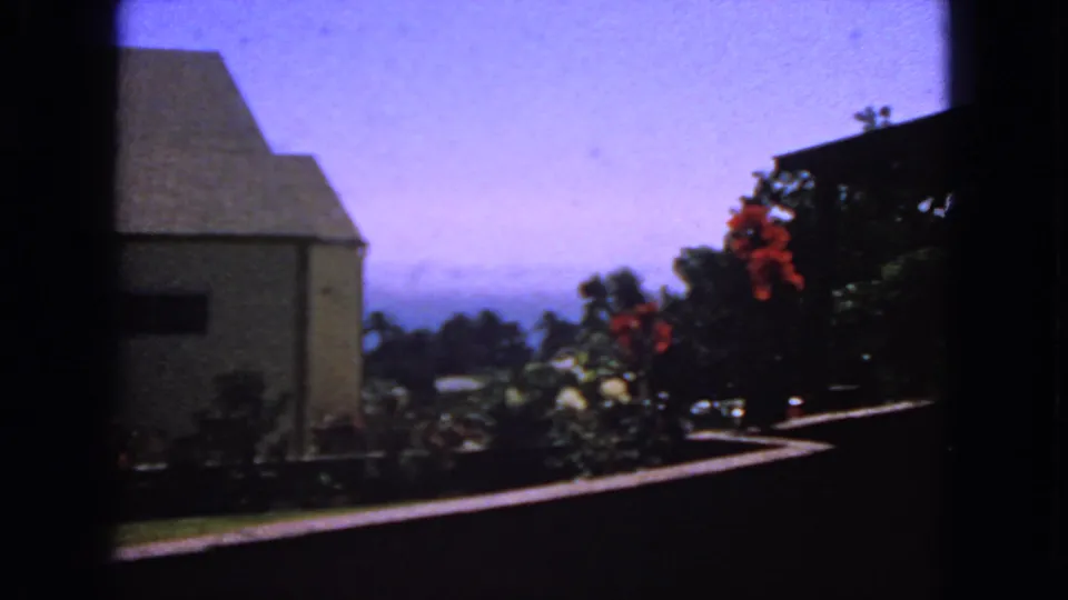 1963 San Francisco frame with a pale sloped-roof house, garden shrubs, red flowers, and hazy blue sky.