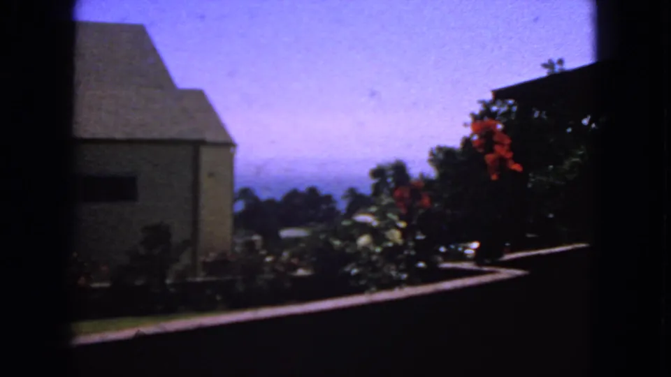 1963 San Francisco home-movie frame showing a residential exterior with garden plants, red blossoms, and distant sky.