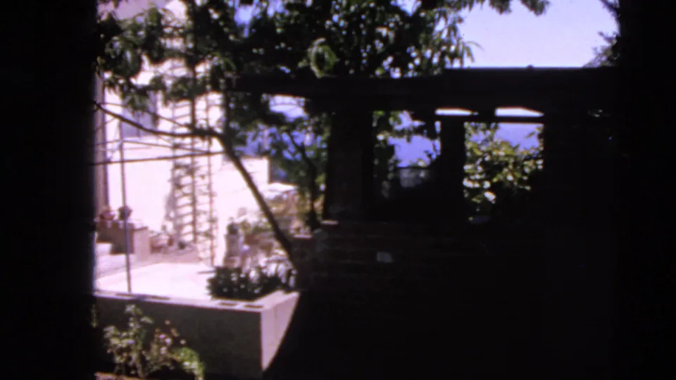 Home-movie view of a San Francisco patio and trellis with trees in the foreground and blue water visible beyond.