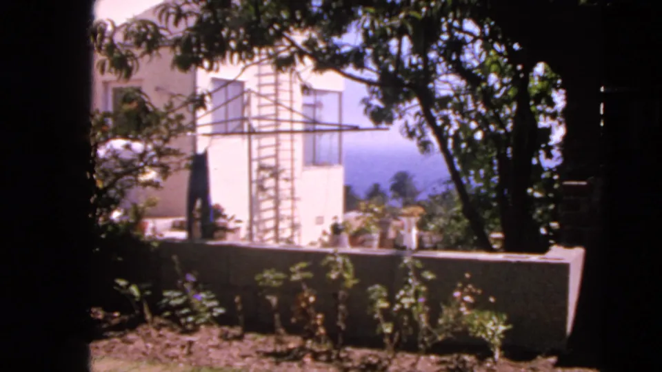 1963 San Francisco 8mm still of a backyard with a white house wall, clothesline structure, trees, garden bed, and low wall.