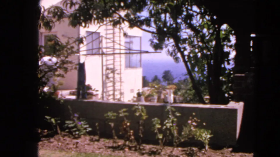 Early-1960s San Francisco home-movie still showing a residential backyard, white exterior wall, clothesline frame, trees, and low wall.