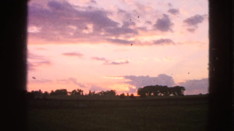 1963 Walworth, Wisconsin 8mm frame showing a pink-orange sunset, low field, distant trees, and dark foreground framing.