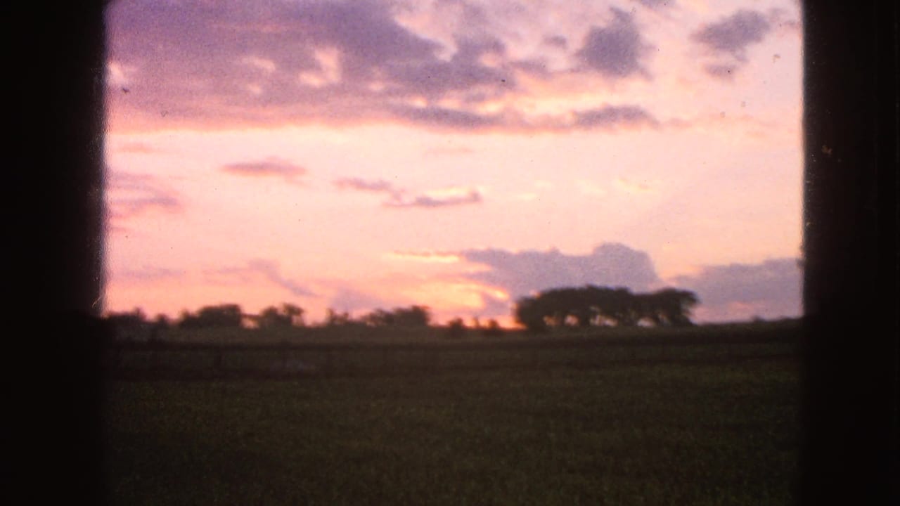 1963 8mm Walworth, Wisconsin view of a pink and lavender sunset over fields and silhouetted trees, seen through dark vertical framing.