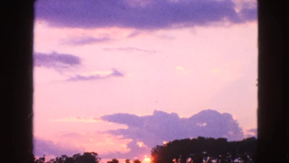1963 Walworth, Wisconsin home-movie frame showing a soft dusk sky, cloud bands, treetops, and a low orange sun glow.