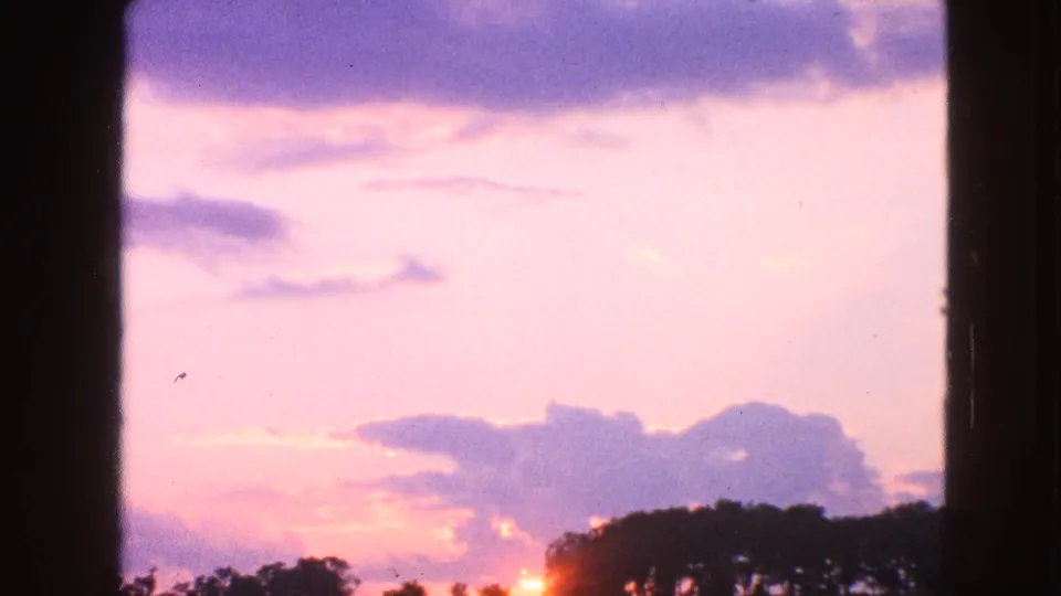 1963 Walworth, Wisconsin 8mm sunset frame with a bright pastel sky, cloud shapes, low sun flare, and dark treetops.