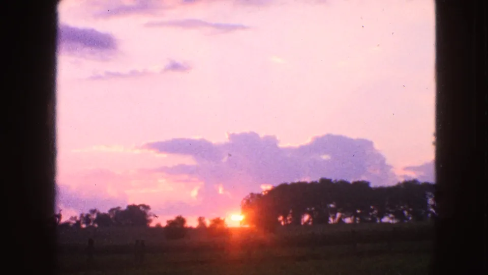 Vintage 8mm view of a bright sunrise flare below purple clouds, with a dark rural treeline and grassy foreground in Walworth, Wisconsin.