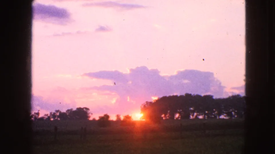 1963 Walworth, Wisconsin 8mm frame showing a bright sunset, silhouetted trees, rural field, and tiny distant human figures near the horizon.