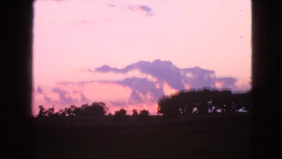 Walworth, Wisconsin home-movie frame from 1963 showing soft pink dusk light over farmland, trees, and purple cloud forms.