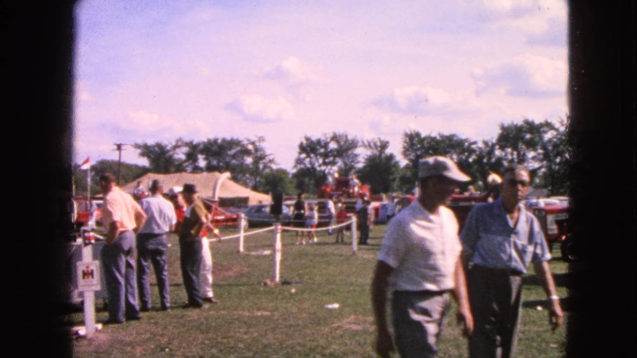 Adult men in 1960s Walworth, Wisconsin home-movie footage walk past roped tractor displays on a sunny grassy field.
