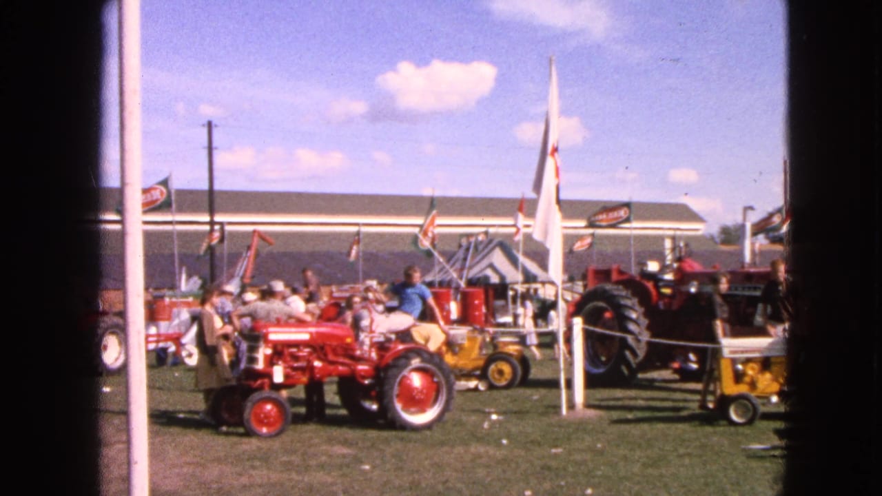 A red tractor and smaller yellow tractor sit near flags and spectators at the 1963 Walworth, Wisconsin tractor show.