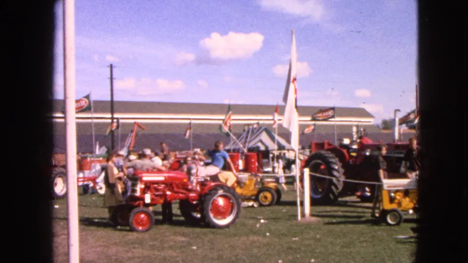 A red tractor and smaller yellow tractor sit near flags and spectators at the 1963 Walworth, Wisconsin tractor show.