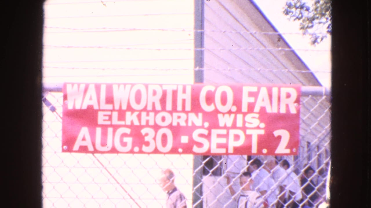 Red Walworth County Fair banner on a chain-link fence with people behind it.