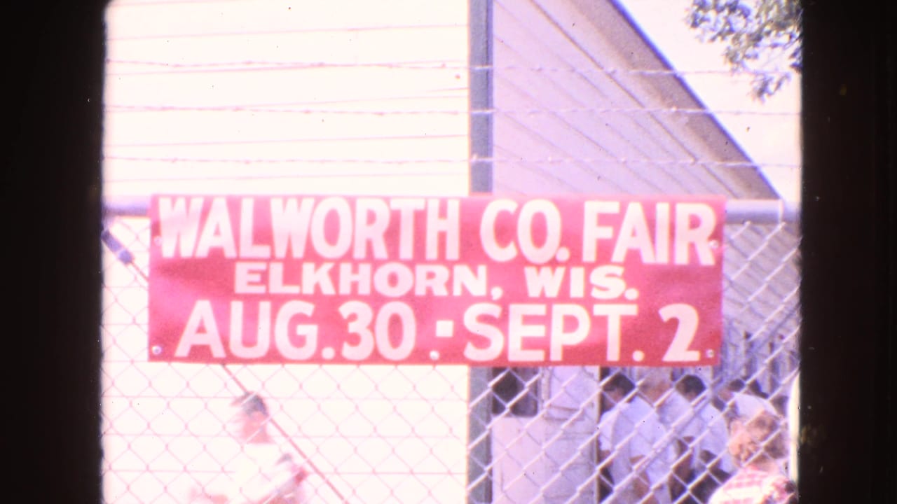 Red Walworth County Fair sign announces August 30 to September 2 while people move near a fairground building.