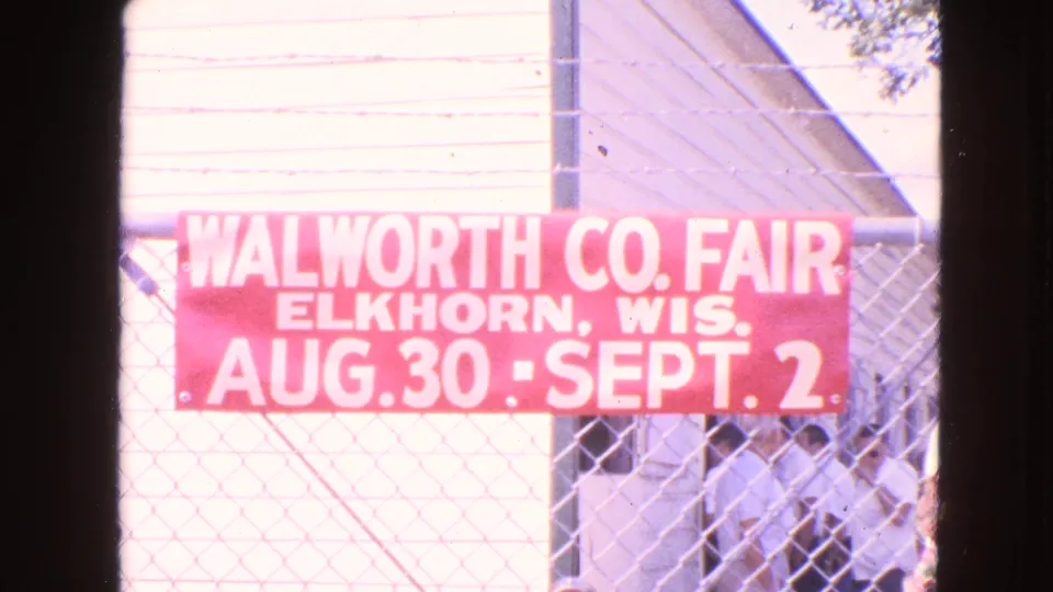 8mm frame centers the Walworth County Fair banner, with a chain-link fence and several visitors near a shaded opening.