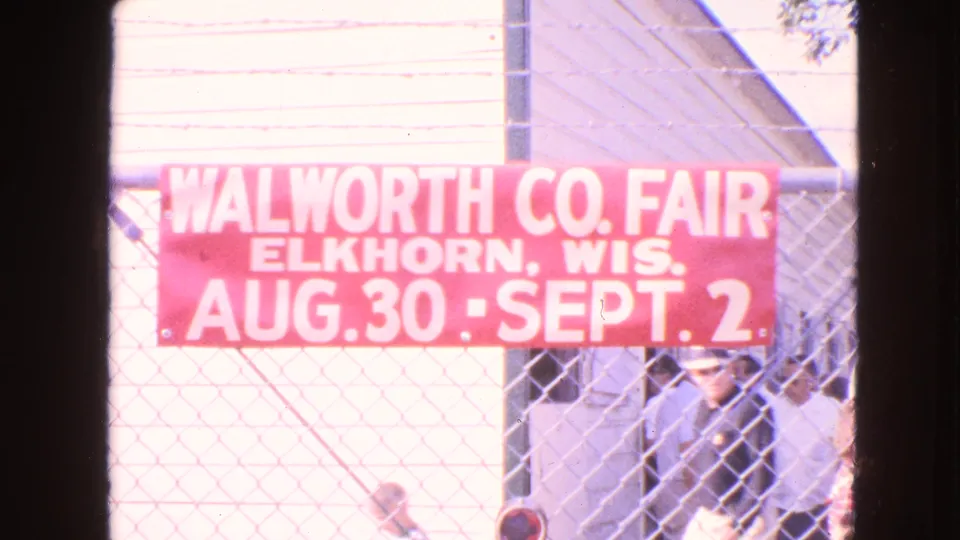 Final frame shows the fair banner and fence, with visitors behind it and one adult face more visible near the lower right.