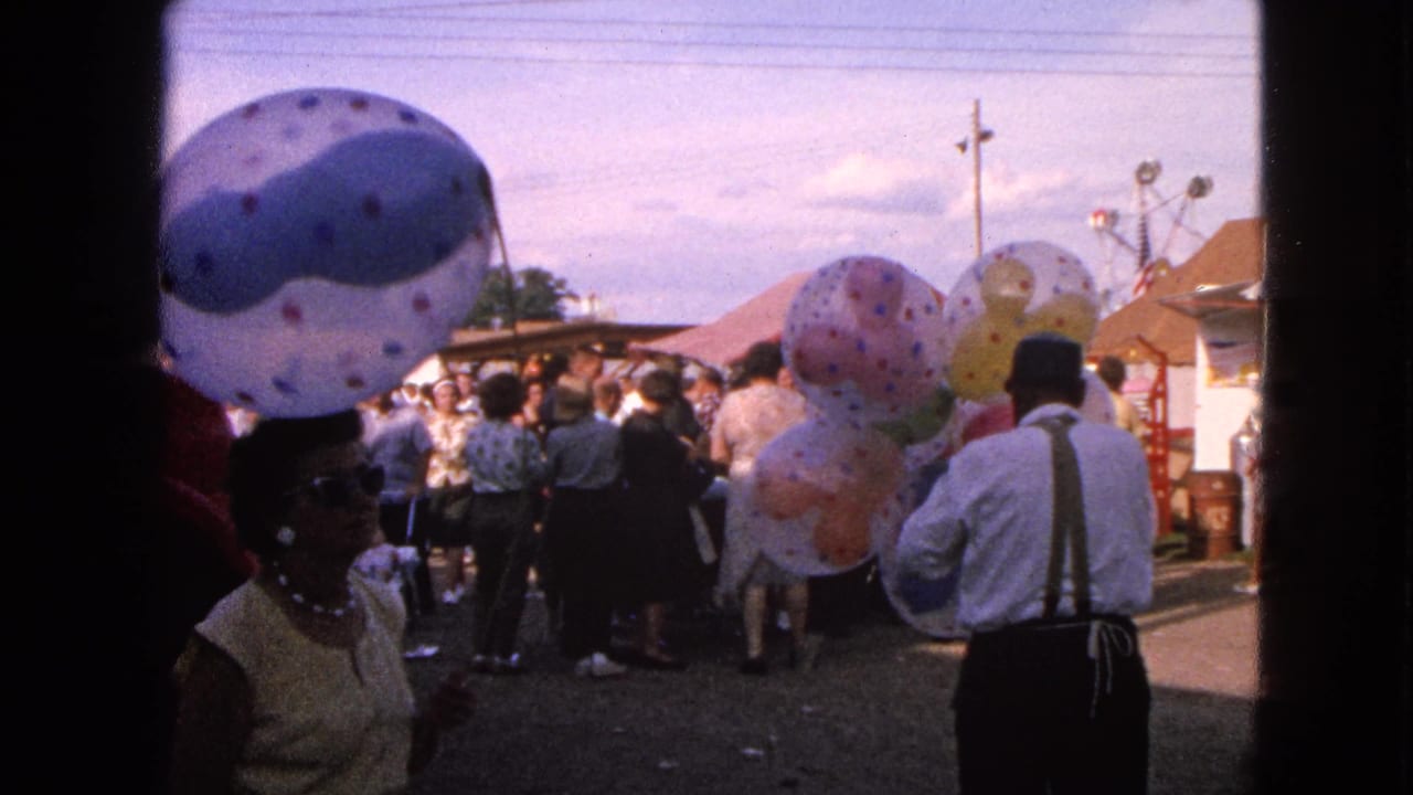 Outdoor crowd with patterned balloons and a man in suspenders near fair tents.