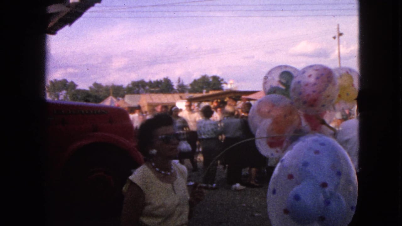 1963 Walworth, Wisconsin frame with a woman in sunglasses near large patterned balloons, a red vehicle edge, and fairgoers in the background.
