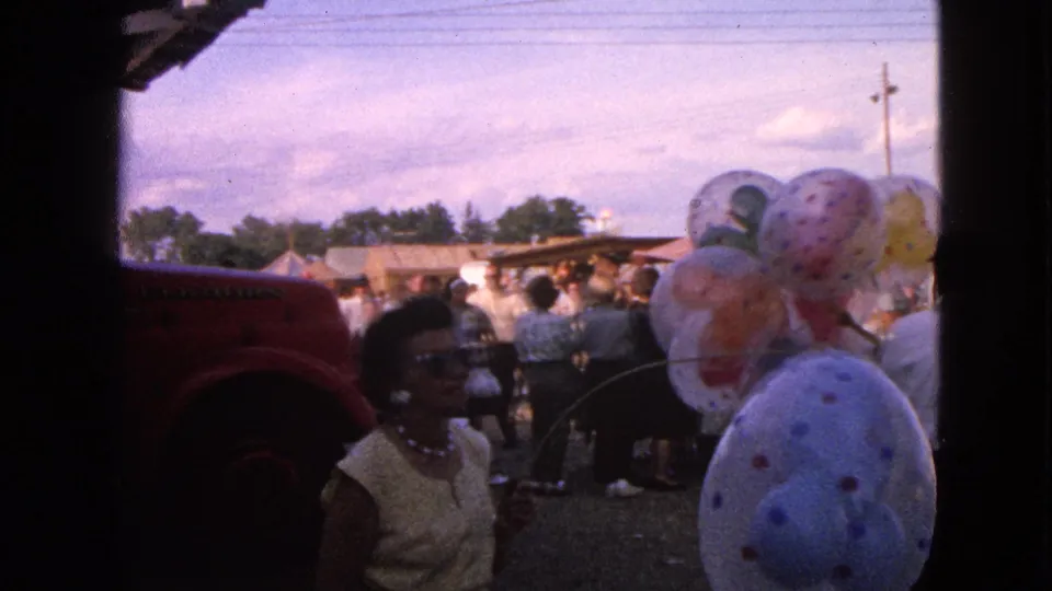 1963 Walworth, Wisconsin frame with a woman in sunglasses near large patterned balloons, a red vehicle edge, and fairgoers in the background.