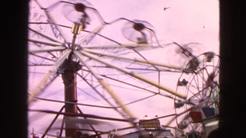 1963 Walworth, Wisconsin 8mm frame showing a Ferris wheel spinning quickly, with riders blurred in open cars and dark film borders at the sides.