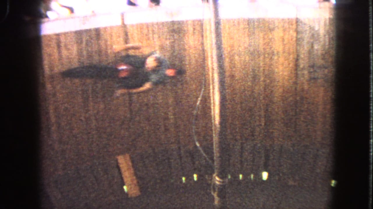 1963 Walworth, Wisconsin 8mm frame showing a blurred rider on a dark racecourse beside a barrier, pole, plank, and green marker lights.