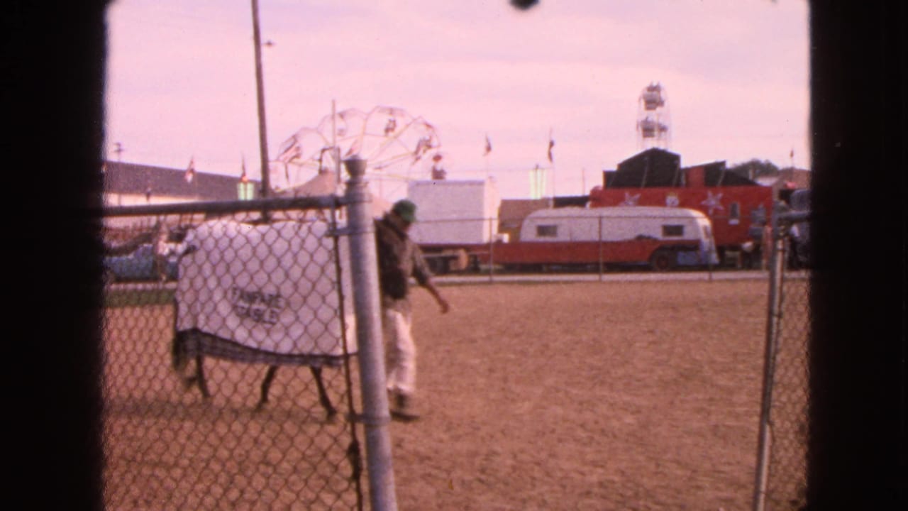 Blanketed horse passing a fence post with a handler in a dirt fairground arena.