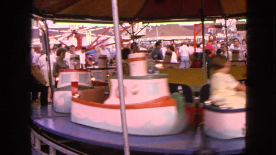 At the 1963 Walworth fairground, a colorful children's boat ride turns beneath a canopy with fairgoers visible around the midway.