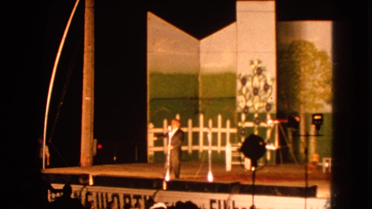 In 1963 Walworth, Wisconsin footage, a suited man stands under warm stage lights before a painted pastoral backdrop and white fence prop.