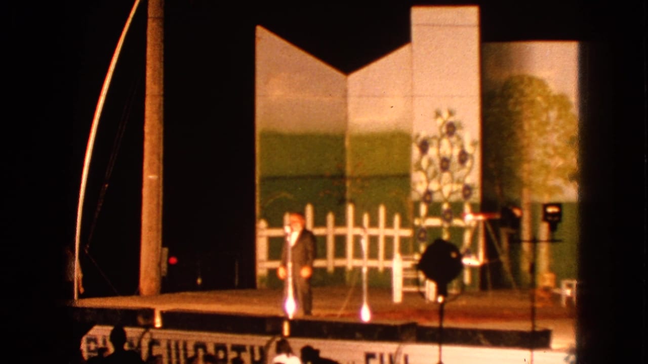 The Walworth stage performance continues as the suited man gestures beside painted flats, a white fence set piece, and visible lighting stands.