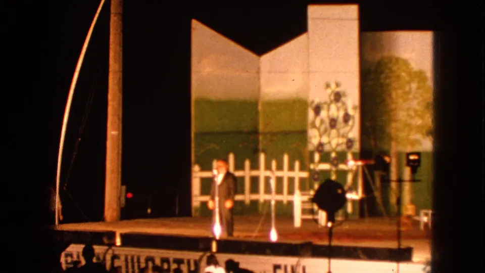The Walworth stage performance continues as the suited man gestures beside painted flats, a white fence set piece, and visible lighting stands.