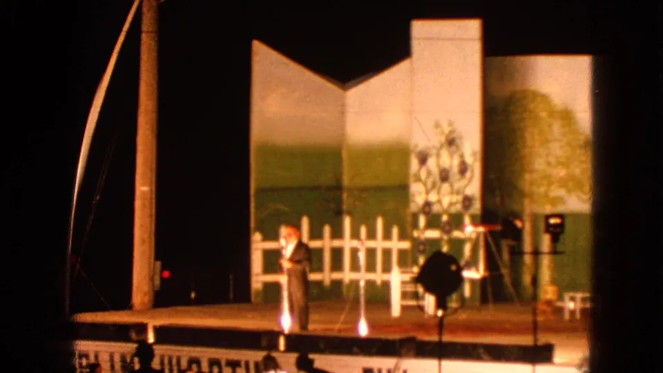 Audience-shot 8mm footage shows the performer near the front of a Walworth stage, with curtains, painted scenery, and audience silhouettes below.