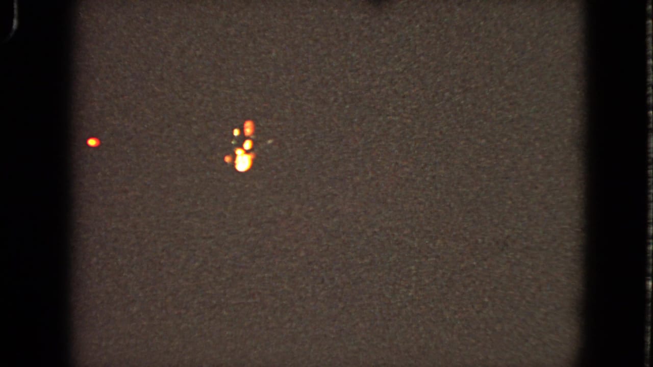 Grainy 1963 home-movie frame from Walworth, Wisconsin showing distant red and orange lights before the stage is visible.