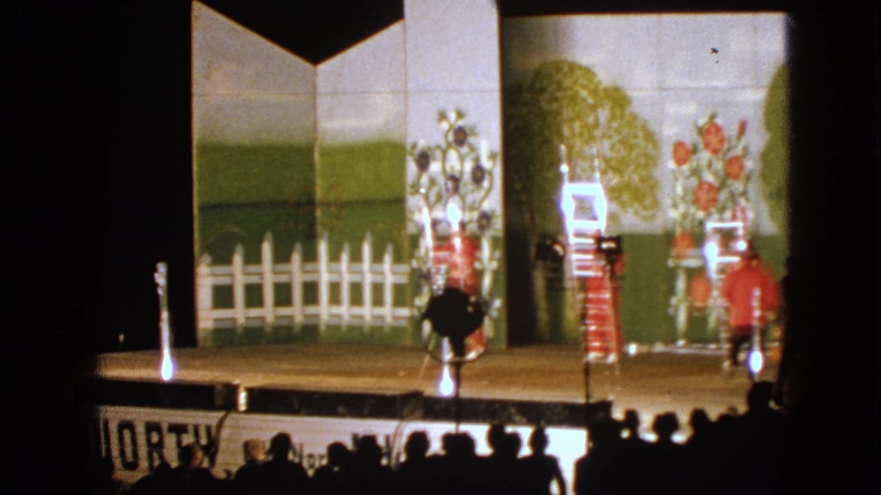 1963 Walworth, Wisconsin home-movie frame showing red-costumed stage performers around ladders before a painted fence, trees, and flowers.