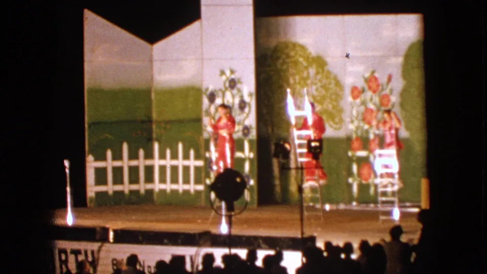 Walworth, Wisconsin 1963 home-movie still of a red-costumed performer near center stage with ladders, lights, and a painted garden backdrop.