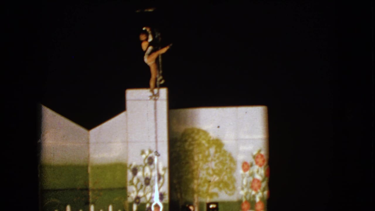 1963 Walworth, Wisconsin 8mm frame showing a woman in costume balancing high on a pole above painted trees and flowers on a dark stage.