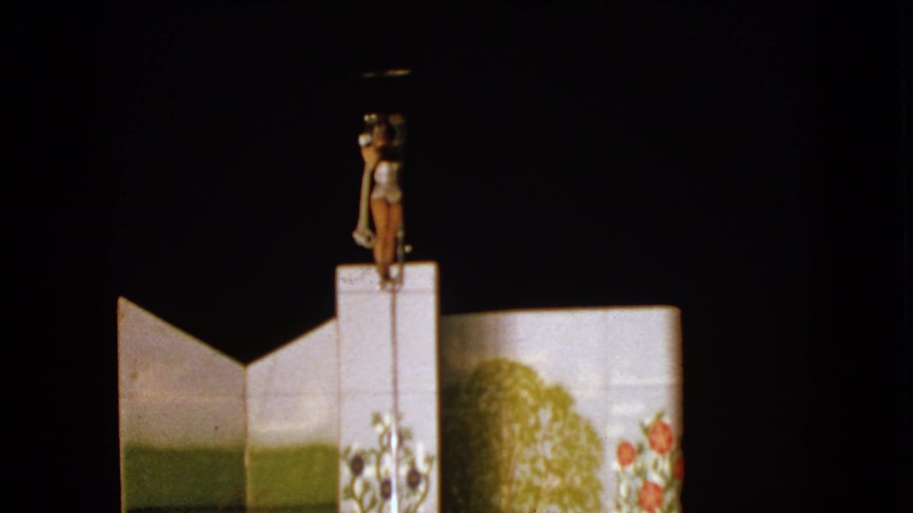 Costumed woman stands on top of a pole set against a dark stage and painted scenery.