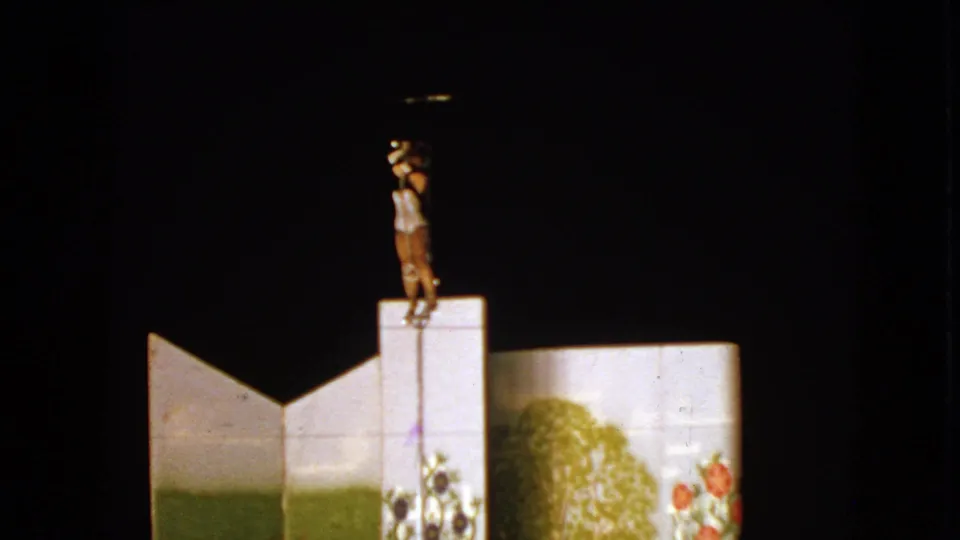 1963 Walworth, Wisconsin 8mm still of a woman balancing on a pole over painted scenery, surrounded by dark theatrical space.