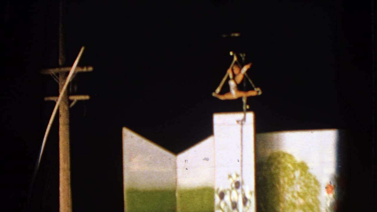 1963 Walworth, Wisconsin 8mm frame showing a trapeze performer suspended above white geometric circus structures under night lighting.