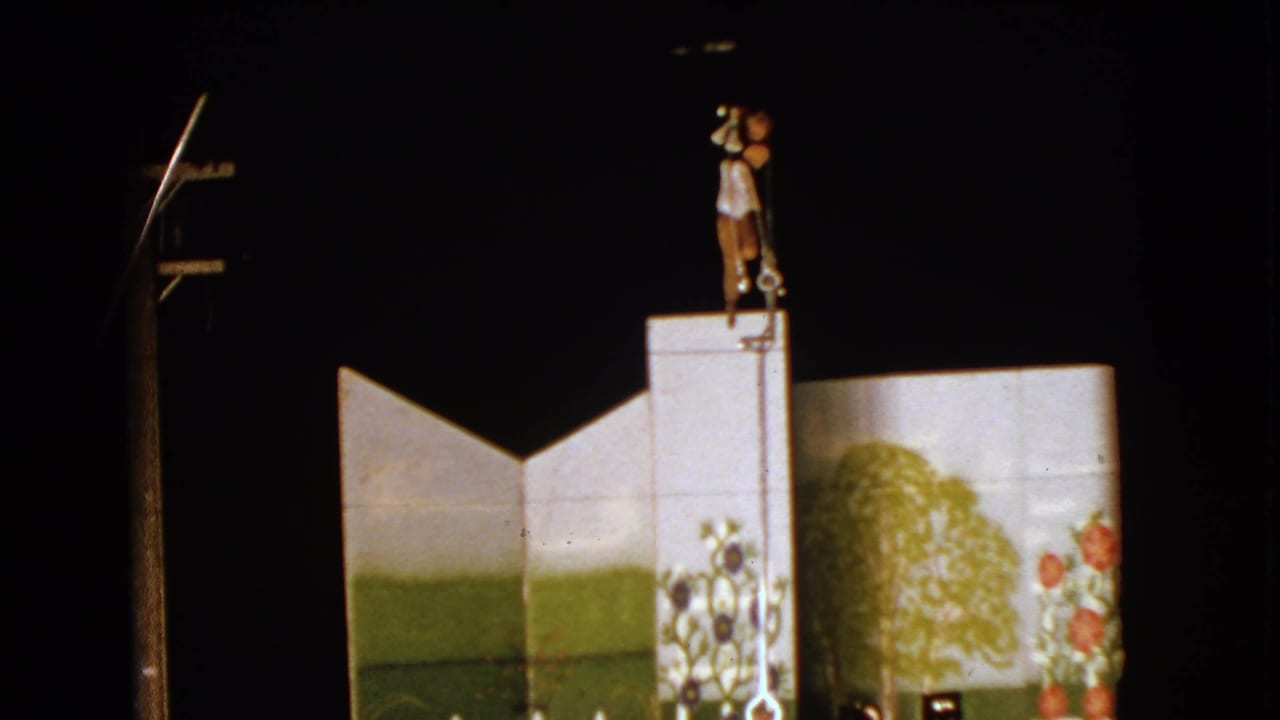 Acrobat stands atop a vertical pole above a painted stage backdrop in a dark circus setting.