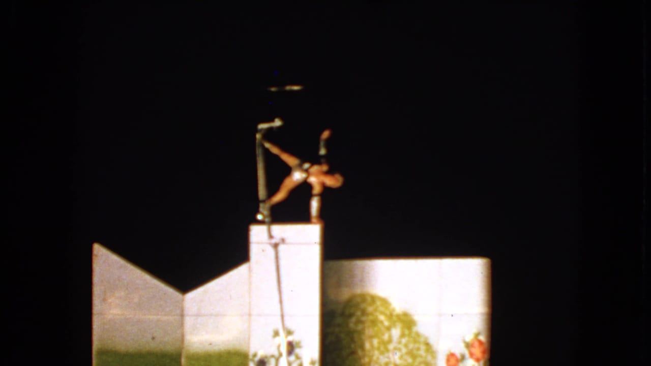 A 1963 circus performer in Walworth extends horizontally from a vertical pole against a dark stage and painted scenery.