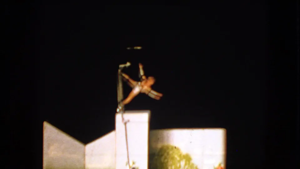 The Walworth 1963 home-movie frame shows the acrobat stretched from the pole above painted grass and flower scenery.