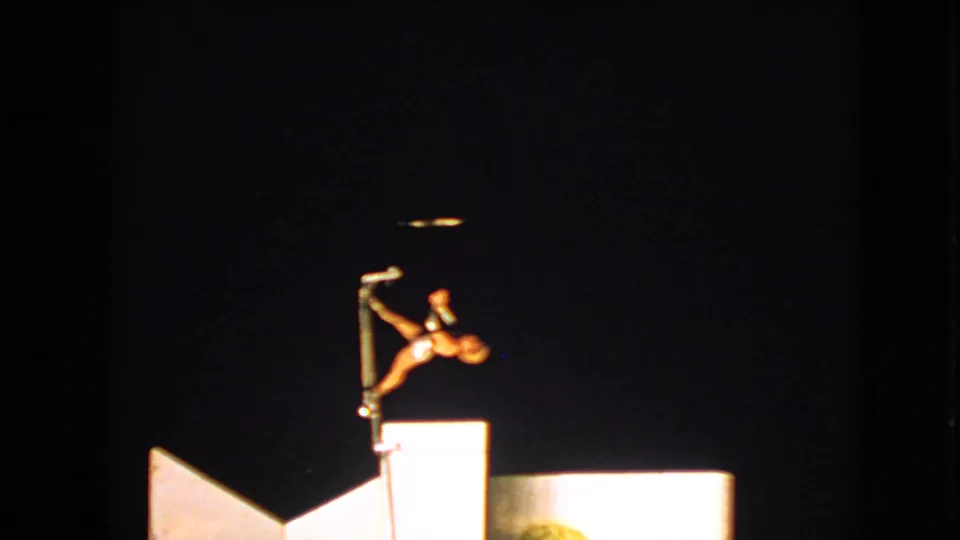 In 1963 Walworth, Wisconsin footage, a female acrobat holds a balancing pose on a pole above a spotlighted platform.
