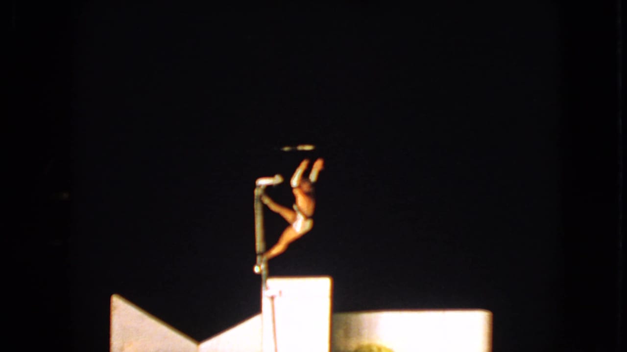 Performer on a pole shifts into an acrobatic pose above a bright stage platform.