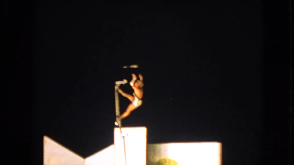 A female performer in the 1963 Walworth footage grips the vertical pole beside a lit platform in a distant audience view.
